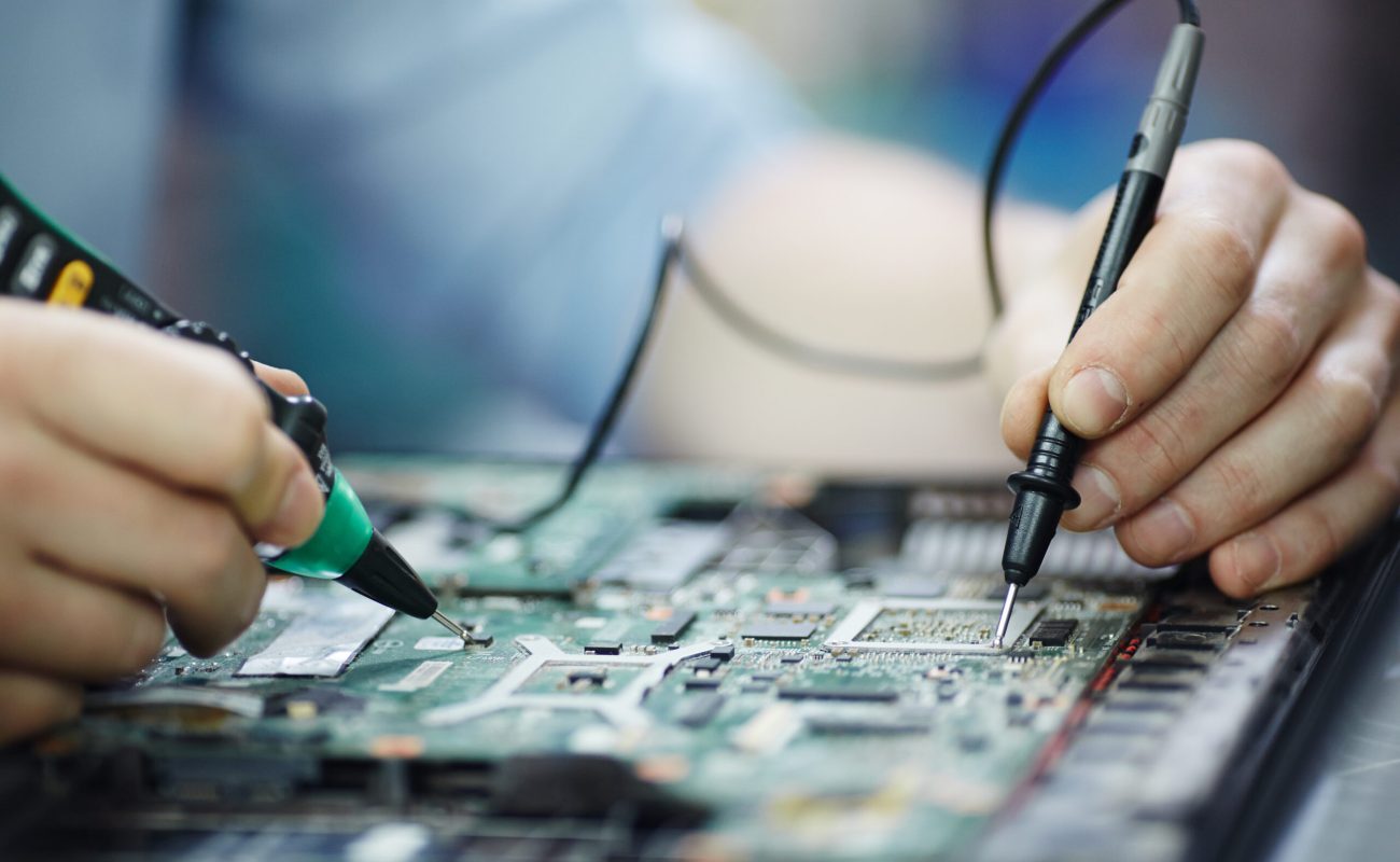 Closeup shot of male hands testing electric current voltage in circuit board of disassembled laptop using multimeter tool on table in maintenance shop